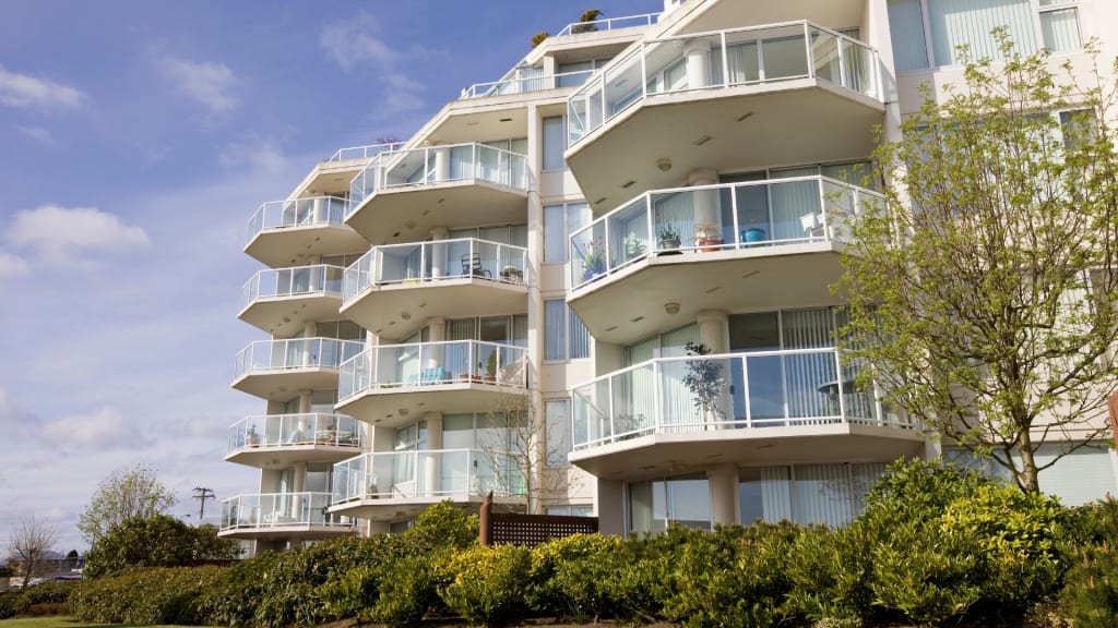 Modern apartment building with multiple glass balconies, white exterior, and surrounding greenery on a clear day.